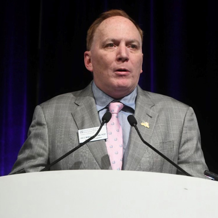Dr. John McManus speaking at a podium wearing gray suit and pink tie.