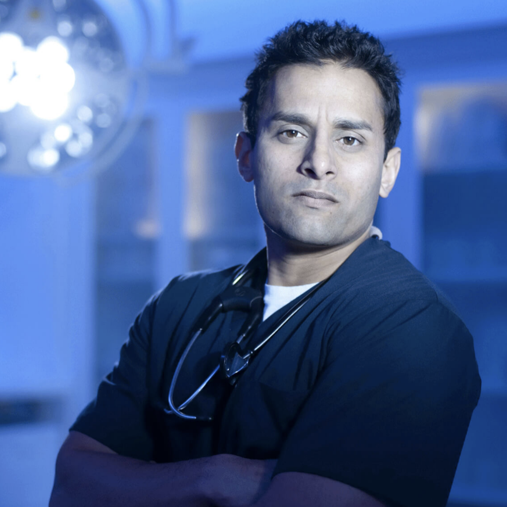 Dr. Sudip Bose standing in surgical theater wearing dark blue scrubs and stethoscope around his neck.